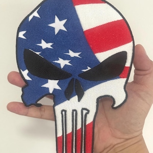 May include: A skull-shaped patch with an American flag design. The top half features the blue field with white stars and red and white stripes. The eye sockets are black, and the teeth are white and red.