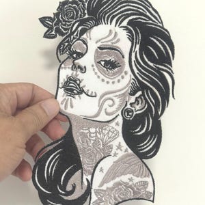 Pode incluir: Patch bordado com uma mulher com maquilhagem do Dia dos Mortos e um adorno floral para o cabelo. O patch é principalmente preto e cinzento, com detalhes intrincados no design facial e tatuagens. O patch é segurado por uma mão.