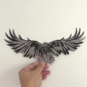 Puede incluir: Un parche bordado en negro y gris con la forma de un pájaro con las alas extendidas. El parche presenta patrones de plumas detallados y se sostiene sobre un fondo blanco.