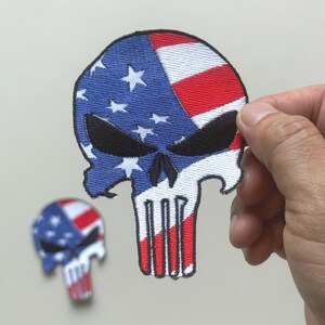 May include: Embroidered patch in the shape of a skull, featuring the American flag design. The skull's top half is blue with white stars, and the other half has red and white stripes. A second, smaller patch is visible in the background.