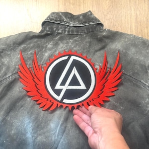 Può includere: Giacca di jeans grigia con una grande toppa ricamata rossa e nera sul retro. La toppa presenta un design circolare con un bordo bianco, un centro nero e un triangolo bianco, con ali rosse che si estendono verso l'esterno.