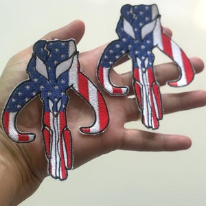 May include: Two embroidered patches featuring a stylized skull design with an American flag motif. The patches have a blue background with white stars, red and white stripes, and white outlines. The patches are held in a hand.