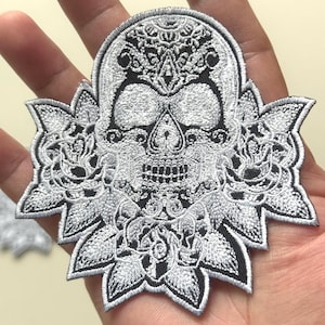 May include: Embroidered patch featuring a detailed skull design with floral accents. The skull is primarily silver with black detailing, surrounded by a stylized floral border. The patch is likely for decorative purposes, suitable for clothing or accessories.