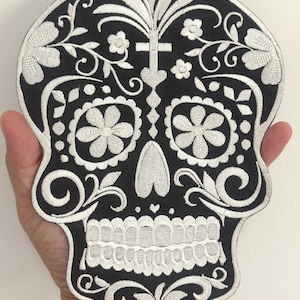 May include: A black and white embroidered patch in the shape of a sugar skull. The design features intricate floral and scrollwork patterns, with a cross at the top. The skull's eyes are represented by large flower shapes.
