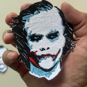 Könnte beinhalten: Gestickter Aufnäher mit einem Porträt einer Figur mit weißem Gesicht, schwarzen Haaren und roten und blauen Akzenten. Der Aufnäher wird in einer Hand gehalten, was die detaillierten Stickereien und das Design zeigt. Für Kleidung oder Accessoires.
