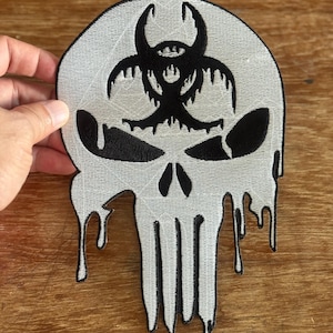 May include: Embroidered patch featuring a skull design with a biohazard symbol on top. The skull is white with black accents, and the design has a dripping effect. The patch is likely for clothing or accessories.