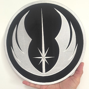 Könnte beinhalten: Ein rundes, schwarz-silbernes Emblem mit einem zentralen Sternenmuster, einer vertikalen Linie und gebogenen flügelartigen Formen. Das Emblem wird in einer Hand gehalten.