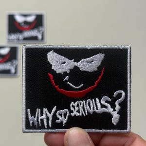Pode incluir: Patch bordado preto com um design de rosto sorridente branco e vermelho e o texto "WHY SO SERIOUS?". O patch tem uma borda branca e é retangular. Outros patches são visíveis no fundo.