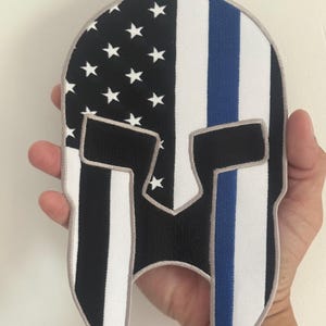 Könnte beinhalten: Gestickter Aufnäher in Form eines spartanischen Helms. Das Design integriert die amerikanische Flagge mit einem blauen Streifen, weißen Sternen auf schwarzem Feld und weißen und schwarzen Streifen.