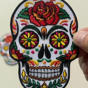 May include: Embroidered skull patch with a white background and black border. The skull features a red rose, colorful floral designs, and a detailed toothy grin. The patch is likely for clothing or accessories.