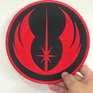 May include: Round black and red embroidered patch featuring a stylized emblem. The design includes a central vertical line, a starburst, and curved wing-like shapes. The patch is held by a hand.