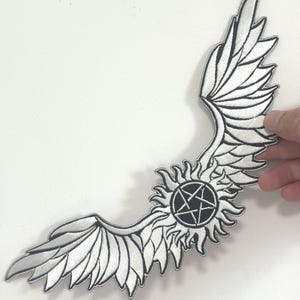May include: Embroidered patch with a wing design, sunburst, and a black pentagram. The patch is white with black detailing. Ideal for customising jackets, bags, or other fabric items. The design is approximately 20 cm in length.