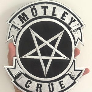 May include: A black and white embroidered patch featuring the words "Mötley Crüe" and a central black circle with a white pentagram. The patch has a distinctive shape with decorative edging.