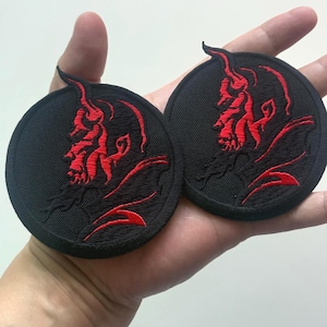 Può includere: Due toppe rotonde nere ricamate con un disegno di diavolo rosso. Il diavolo ha corna e barba, con il disegno centrato sulla toppa. Le toppe sono tenute in una mano.