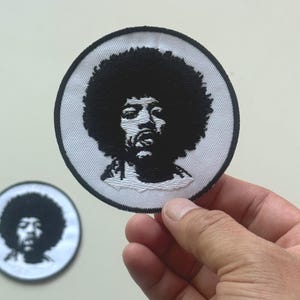 Puede incluir: Un parche redondo en blanco y negro con un retrato de una persona con afro. El parche tiene un fondo blanco con un borde negro. Un segundo parche es visible en el fondo.