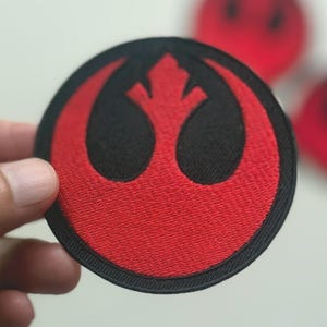 Könnte beinhalten: Runder, gestickter Aufnäher mit dem Symbol der Rebellenallianz aus Star Wars. Der Aufnäher hat einen schwarzen Rand und ein rotes Emblem. Das Design ist eine stilisierte Vogel- oder Phönixform. Weitere Aufnäher sind im Hintergrund sichtbar.