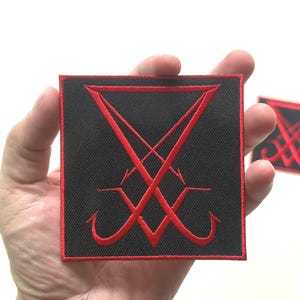 May include: Black square patch with a red sigil design. The patch has a red border and is held in a hand. The sigil is a stylized symbol with inverted V shapes and curved lines.