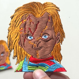 May include: Embroidered patch featuring a stylized Chucky doll face with orange hair, blue eyes, and stitched scars. The patch has a brown face, and a colorful shirt with red, blue, and green accents. The patch is likely for clothing or accessories.