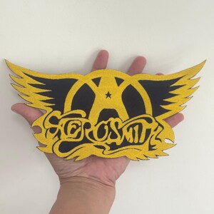 May include: Aerosmith logo patch in black and gold. The patch features a stylized winged design with the band's name in a decorative font. The patch is held in a hand.