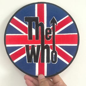 Può includere: Toppa circolare con il design della bandiera Union Jack in rosso, bianco e blu. Il testo nero "The Who" ha una freccia sopra la lettera "e". La toppa è tenuta da una mano.