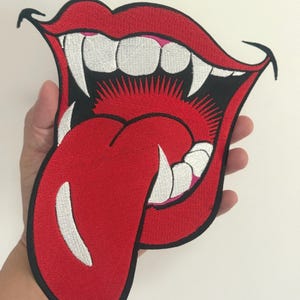 May include: Embroidered patch featuring a red mouth with a long, red tongue sticking out. The mouth has white teeth and fangs, with a black outline. The patch is likely for clothing or accessories.