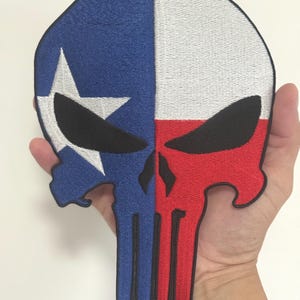 May include: Embroidered patch in the shape of a skull, split vertically. The left side is blue with a white star, the right side is red and white. The skull has black eye sockets and a black outline.