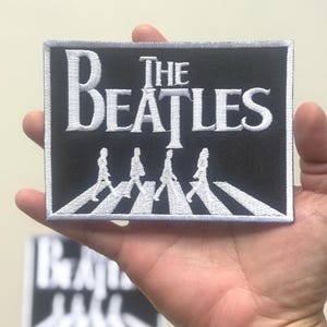 Könnte beinhalten: Ein rechteckiger, schwarz-weißer, gestickter Aufnäher mit dem Text "THE BEATLES" in weißer, stilisierter Schrift. Darunter eine weiße Darstellung der Bandmitglieder, die über einen Zebrastreifen gehen. Der Aufnäher hat einen weißen Rand.