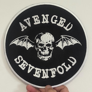 May include: A round, black patch with white embroidered text and a skull graphic. The text reads "AVENGED SEVENFOLD" in a curved font around the skull with bat wings. The patch is likely for a jacket or bag.