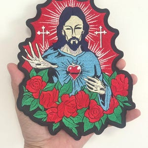 Puede incluir: Parche bordado que representa una figura religiosa con un corazón rojo, rodeada de rosas rojas y un halo rojo. La figura tiene pelo largo y oscuro y barba, vestida con una túnica azul. Dos cruces blancas son visibles.