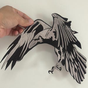 May include: Black and gray embroidered patch in the shape of a bird with outstretched wings. The patch features detailed stitching, with the bird's feathers and body outlined in black and filled with gray.