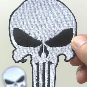 May include: White and black embroidered skull patch with a stern expression. The patch features a detailed design with black eye sockets and a vertical line pattern for the teeth. A smaller, similar skull emblem is visible in the background.