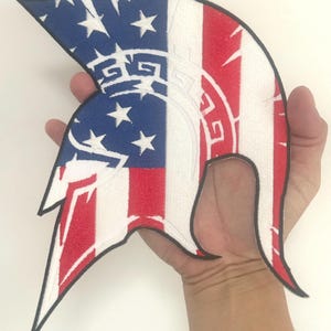 Op de afbeelding: Een patriottische patch in de vorm van een Spartaanse helm. Het ontwerp toont de Amerikaanse vlag met rode en witte strepen en een blauw veld met witte sterren. De helm heeft een zwarte omtrek.