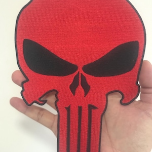 May include: A red skull patch with black eye sockets and a black nose and mouth. The patch is made of fabric and has a stitched design. The skull has a distinctive shape, with a prominent forehead and jawline.