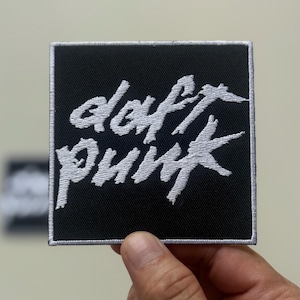 Puede incluir: Un parche cuadrado negro con texto bordado blanco "daft punk". El parche tiene un borde blanco y está sostenido por una mano. Este parche temático musical es para ropa o accesorios.