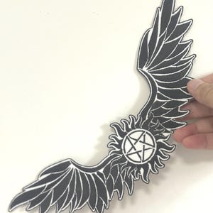 Può includere: Toppa in bianco e nero con un design stilizzato di ali d'angelo. La toppa include un emblema centrale a forma di sole con una stella a cinque punte. Le ali sono dettagliate con contorni bianchi.