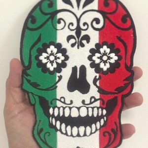 May include: Embroidered skull patch with a design featuring green, white, and red stripes, floral accents, and a black outline. The skull has a wide grin and flower-shaped eye details. This patch is suitable for jackets, bags, or other fabric items.