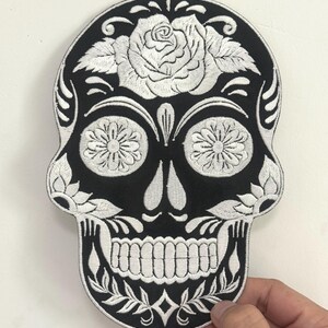 May include: Black and white embroidered skull patch. The patch features a detailed design with a rose at the top, floral accents, and a grinning skull. The patch is held by a hand.