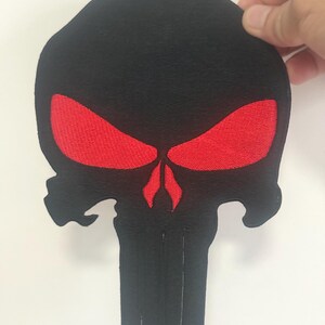 May include: Black skull patch with red eyes and a red nose. The patch is held by a hand. The skull design is a popular symbol often associated with comics, movies, and gaming.