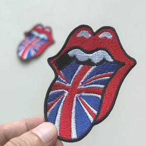 May include: Embroidered patch featuring the iconic Rolling Stones tongue and lips logo. The design incorporates the Union Jack flag within the tongue, with red lips and a black outline. A smaller, similar patch is visible in the background.