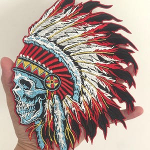 May include: Embroidered patch featuring a Native American headdress design with a skull. The patch is predominantly red, black, white, and blue, with intricate feather and skull details. The patch is likely for clothing or accessories.