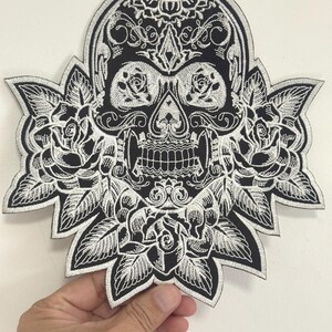 May include: Black and white embroidered patch featuring a sugar skull design with floral accents. The skull has detailed facial features and is surrounded by roses and leaves. The patch is held against a white background.