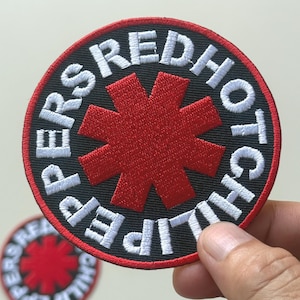 May include: Embroidered patch featuring the text "Red Hot Chili Peppers" in white, encircling a red asterisk on a black background. The patch has a red border and is held by a hand. Another patch is visible in the background.
