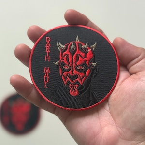May include: A circular, black embroidered patch depicting Darth Maul from Star Wars. The patch features a red and black design, with the character's face and the text "Darth Maul" in red thread. Another similar patch is visible in the background.