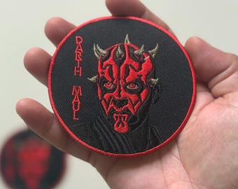 Può includere: Una toppa rotonda e nera ricamata con il personaggio di Darth Maul di Star Wars. La toppa ha una combinazione di colori rosso e nero, con il volto del personaggio e la scritta "Darth Maul" in filo rosso. Una seconda toppa simile è visibile sullo sfondo.