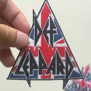 Puede incluir: Un parche triangular con el texto "DEF LEPPARD" en negro, sobre un diseño de la bandera británica en rojo, blanco y azul. El parche tiene un borde blanco.