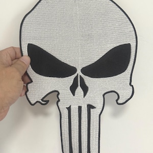 May include: A gray and black skull patch with a detailed embroidered design. The skull features black eye sockets, a nose, and a jawline. The patch is held by a hand against a white background.