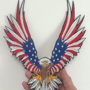 May include: Embroidered patch depicting an eagle with wings spread wide. The design uses the colours of the American flag: red and white stripes, a blue section with white stars. The eagle's body is brown, with yellow talons and white feathers.