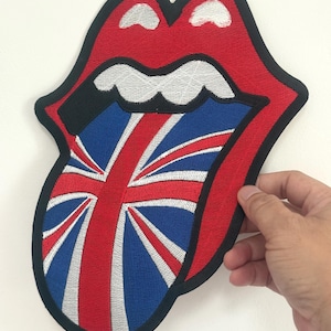 May include: A red, black, and white embroidered patch in the shape of a mouth with a tongue. The tongue features the Union Jack flag design in blue, red, and white. The patch has a black border.