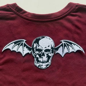 Puede incluir: Una camiseta burdeos de cuello redondo con un parche blanco y negro. El parche representa una calavera con alas de murciélago. La calavera tiene sombreado detallado y las alas tienen una apariencia texturizada. La tela de la camiseta tiene un aspecto suave y ligeramente texturizado.