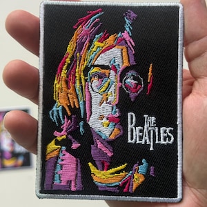 Könnte beinhalten: Gestickter Aufnäher mit einem farbenfrohen, abstrakten Porträt einer Person und dem Text "The Beatles" in Weiß. Der Aufnäher hat einen schwarzen Hintergrund und einen weißen Rand.